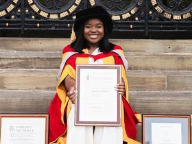 Vee Kativhu wearing academic robes holding up her three degree certificates
