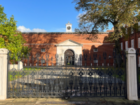 A beautiful image of the front of Lady Margaret Hall, with a set of wrought iron gates in front of an imposing red brick building set against a bright blue sky