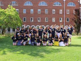 A large group of students in navy t-shirts stand on bright green grass waving at the camera