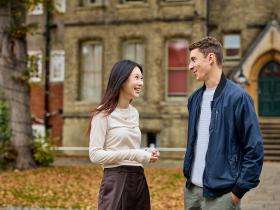 Two students, one male and one female, stand talking animatedly outside of a victorian-style brick building