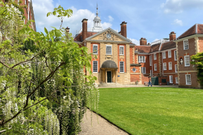 In the foreground, white wisteria is growing up the wall of the building. Further away is an imposing red brick building set in a quad