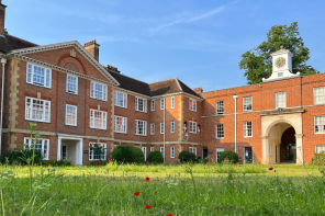 Red brick buildings, one with a clock tower, and in the foreground bright green grass with pops of red poppies