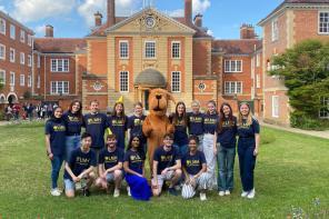 A group of smiling students wearing matching navy blue LMH (Lady Margaret Hall) t-shirts pose in front of a grand brick college building with a domed entrance. A person in a large brown dog mascot costume stands at the center. The group is gathered on a green lawn under a sunny sky with fluffy clouds.