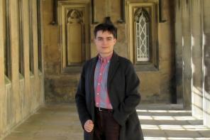 A young man in a long black coat standing in some stone cloisters