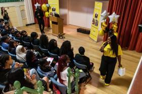 A group of young people in a lecture theatre, with Vee Kathivu wearing a yellow tshirt and holding a microphone