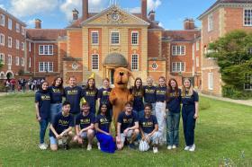 A group of smiling students wearing matching navy blue LMH (Lady Margaret Hall) t-shirts pose in front of a grand brick college building with a domed entrance. A person in a large brown dog mascot costume stands at the center. The group is gathered on a green lawn under a sunny sky with fluffy clouds.