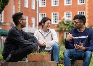 Three students, two men of Asian descent and a white woman, sit outside a red brick building talking and laughing