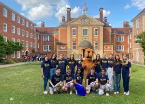 A group of smiling students wearing matching navy blue LMH (Lady Margaret Hall) t-shirts pose in front of a grand brick college building with a domed entrance. A person in a large brown dog mascot costume stands at the center. The group is gathered on a green lawn under a sunny sky with fluffy clouds.
