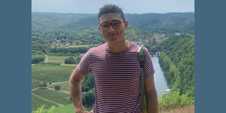 Adam Husain, who has short dark hair and glasses and is wearing a red and white striped t-shirt standing with a beautiful countryside landscape in the background