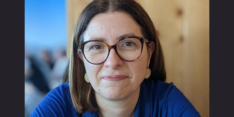 Maria Coelho, who has short dark brown hair and glasses and is wearing a blue top, smiles at the camera