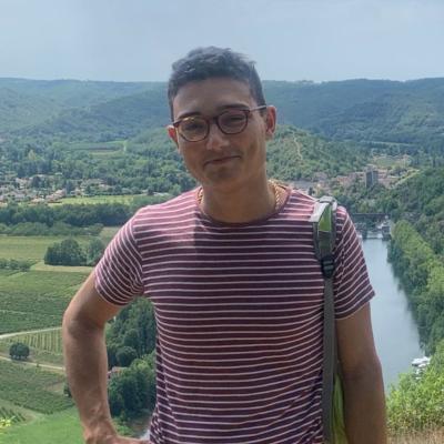 Adam Husain, who has short dark hair and glasses and is wearing a red and white striped t-shirt standing with a beautiful countryside landscape in the background