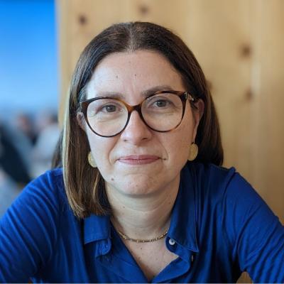 Maria Coelho, who has short dark brown hair and glasses and is wearing a blue top, smiles at the camera