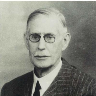 A black and white portrait of Jules Strauss, an elderly man with small oval framed glasses and a grey moustache
