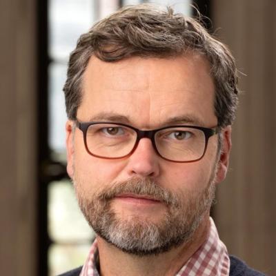 A man with short brown hair and a beard with rectangular-framed glasses looks at the camer